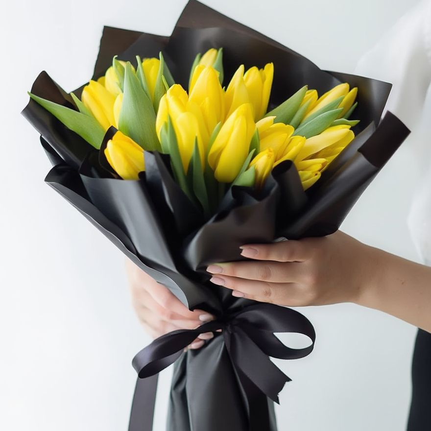 Bouquet of yellow tulips wrapped in black paper with a hand holding it against a white background