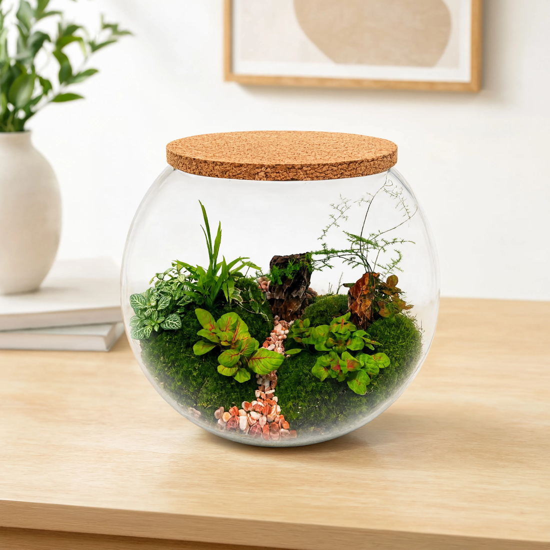 Handcrafted round glass terrarium with live moss, tropical plants, decorative driftwood, and natural pebble pathway displayed on a wooden table in a modern living room.