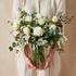 Person holding a bouquet of white roses and greenery in a clear vase against a beige background