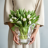 Person holding a clear vase with white tulips against a neutral background