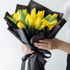 Bouquet of yellow tulips wrapped in black paper with a hand holding it against a white background